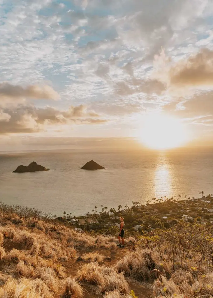 Me standing on the top of the Lanikai Pillbox trail overlooking the ocean at sunrise. The sky is filled with clouds, and the sun is rising over two small islands in the distance. Below, a town with scattered palm trees is visible along the shoreline.
