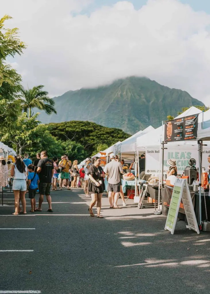 People walking to stands at a farmers market in Kailua Oahu with green mountains and foliage in the distance