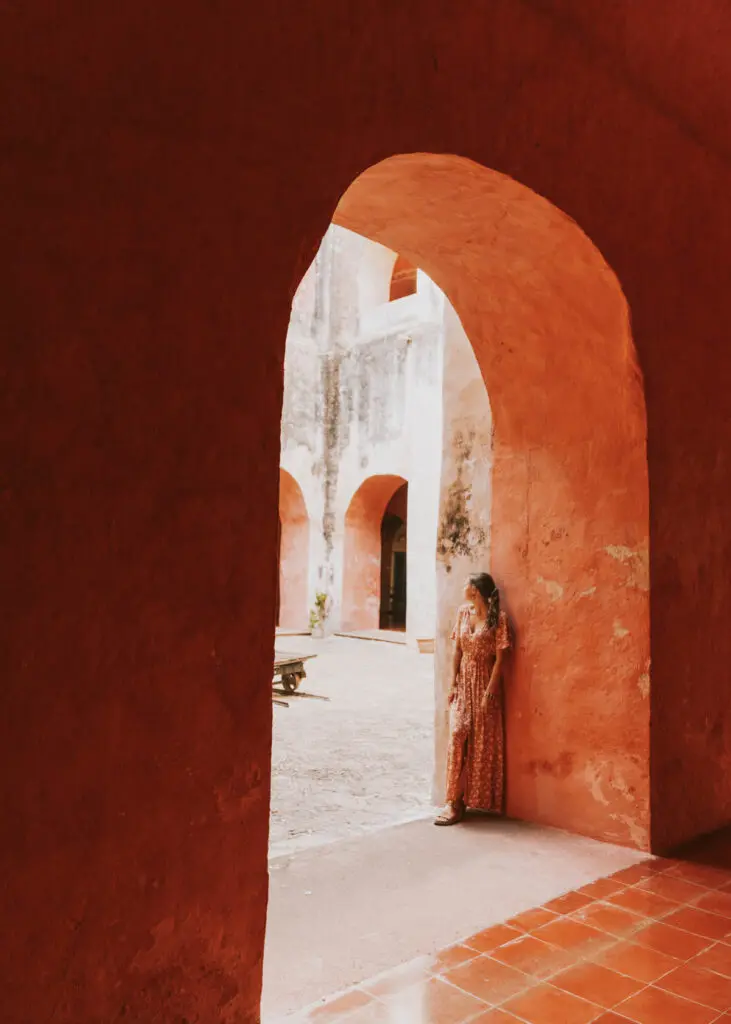 A woman in a long dress leans against the red stone archway of a historical building in Valladolid Mexico. The architecture features rustic textures and warm earth tones, with additional arches in the background.