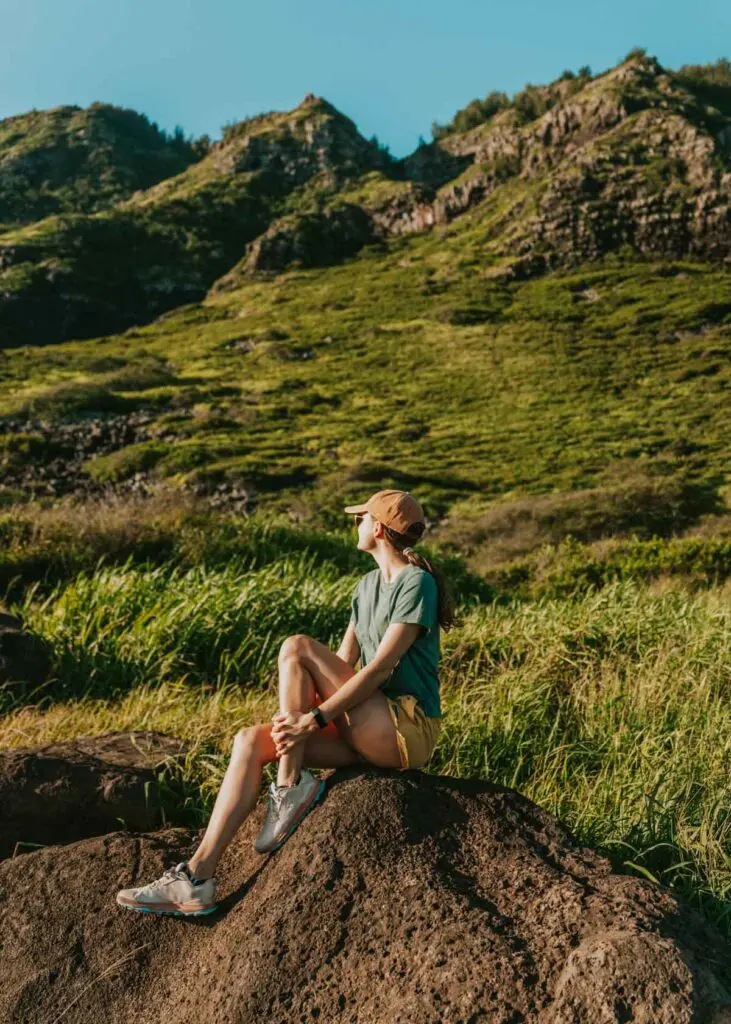 Woman sitting on rock in front of mountains at the Kaena Point State Park parking lot on Oahu's North Shore.