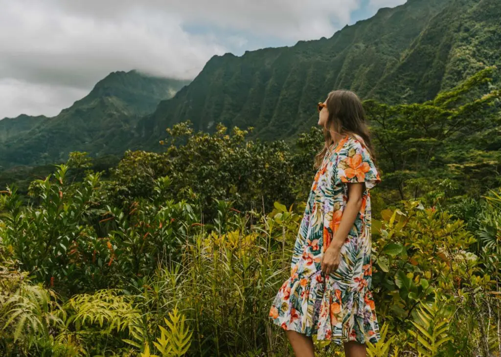 A woman in a floral dress stands in front of green ridged mountains and tropical foliage in a botanical garden on Oahu, Hawaii