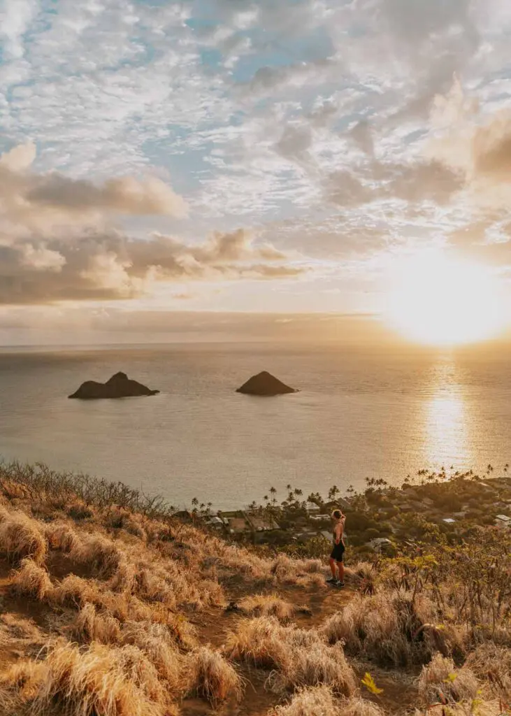 A person stands on a grassy mountain peak overloooking the ocean at sunset. Two small islands are visible in the distance under a partly cloudy sky. The sun casts a warm glow over the scene.