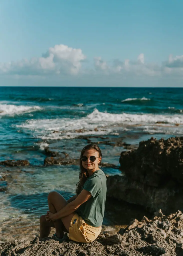 A person with long hair sits on a rocky shore in Haleiwa, wearing sunglasses, a green shirt, and yellow shorts. Behind them on Oahu's stunning beaches, gentle waves roll in under a blue sky with scattered clouds.