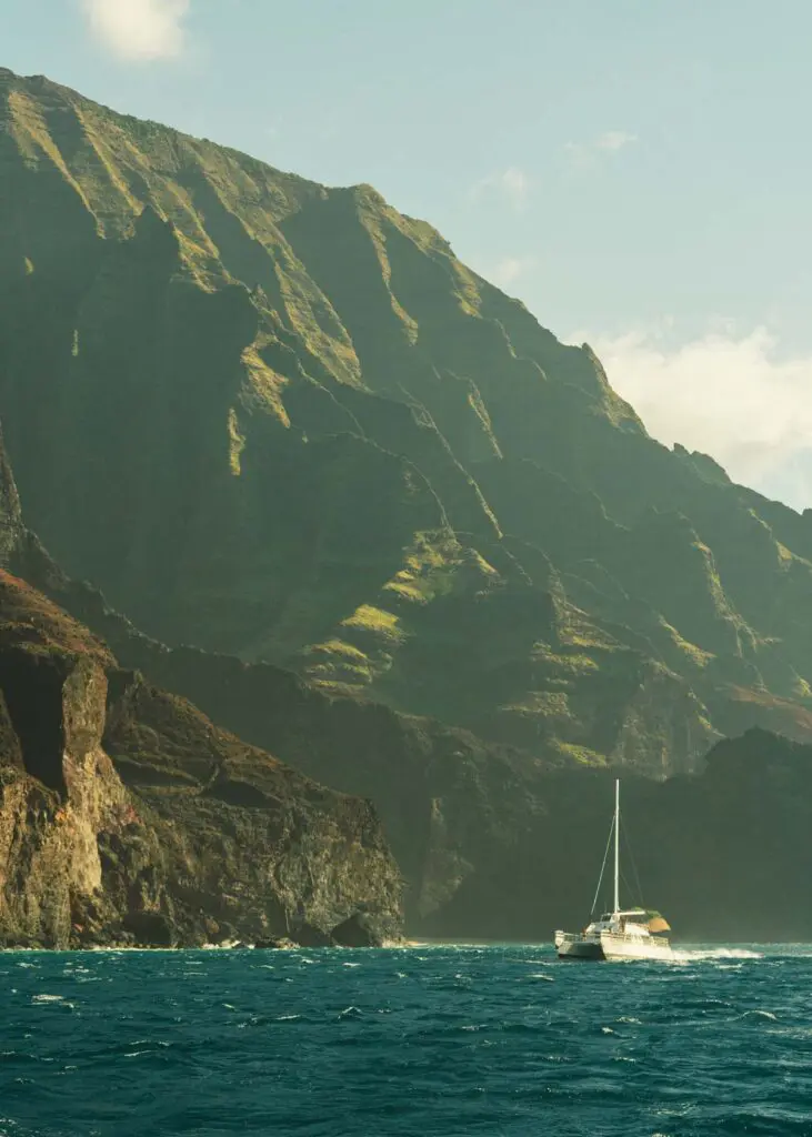 A catamaran sailboat riding the waves along the Napali Coast in Kauai with verdant cliffs overlooking the choppy sea