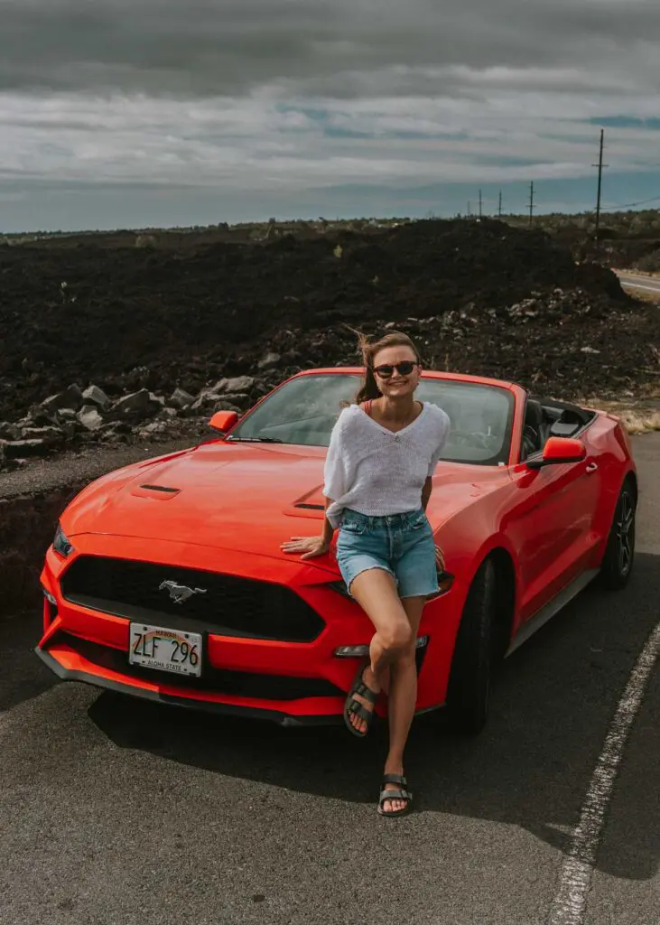 Leaning on the front of a red Mustang convertible rental car on the Big Island of Hawaii. There are lava fields in the background.