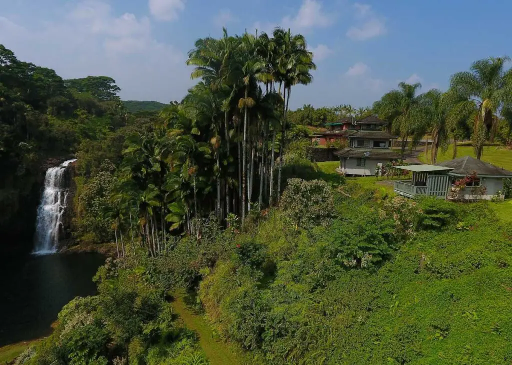 A lush green landscape on the Big Island features a waterfall cascading into a pool on the left, surrounded by tall palm trees. A house with a spacious gazebo sits among the greenery on the right under Hawai'i's clear blue sky.