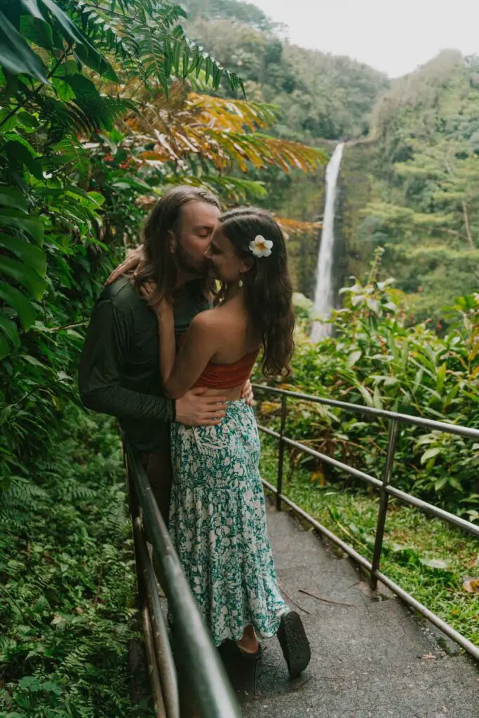 A couple embraces lovingly on a narrow path surrounded by lush greenery on the Big Island. A waterfall is visible in the background. The woman wears a floral skirt and a flower in her hair, while the man has long hair and a beard, capturing the spirit of Hawai'i's natural allure.