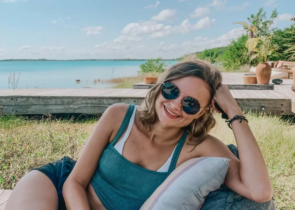 A woman in sunglasses and a blue tank top relaxes on a cushioned seat by Bacalar lagoon at Our Habitas Bacalar.