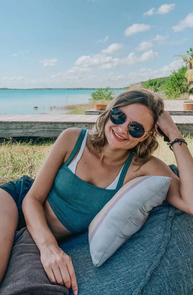 A woman in sunglasses and a blue tank top relaxes on a cushioned seat by Bacalar lagoon at Our Habitas Bacalar.