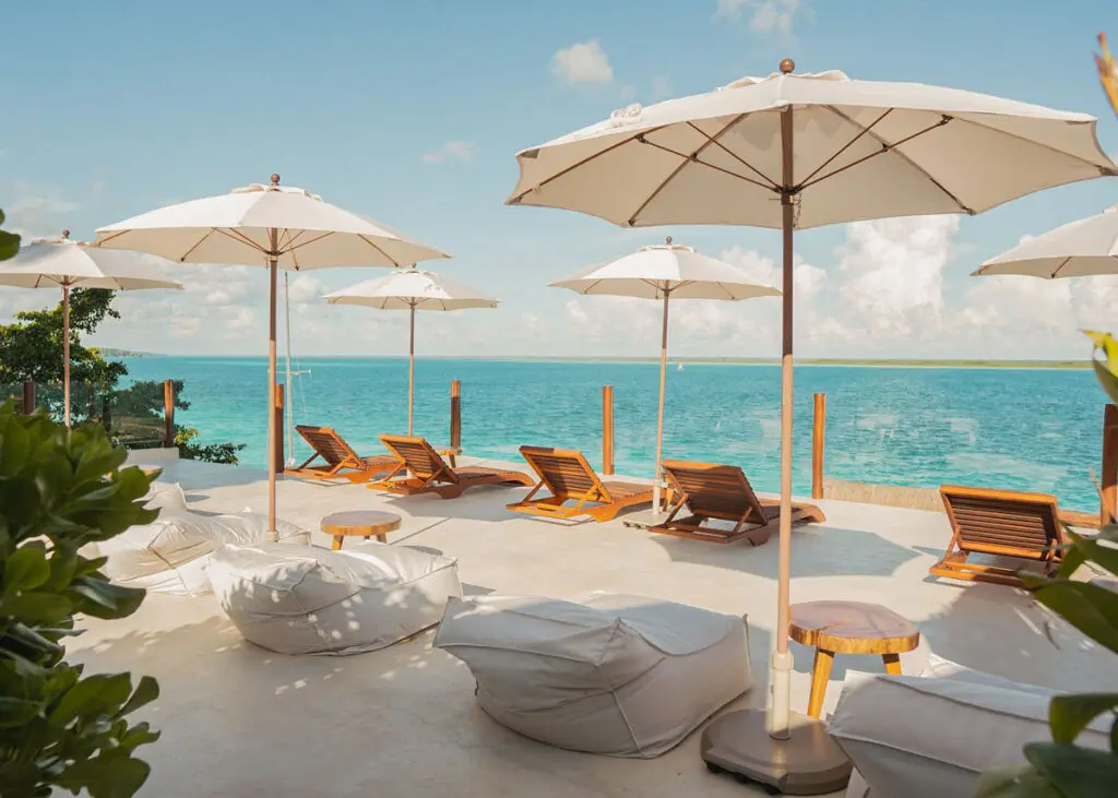 Wooden lounge chairs and white sun loungers with white umbrellas line a cement deck overlooking Bacalar lagoon at Puerta del Cielo Hotel Origen, Bacalar's highest rated waterfront hotel.