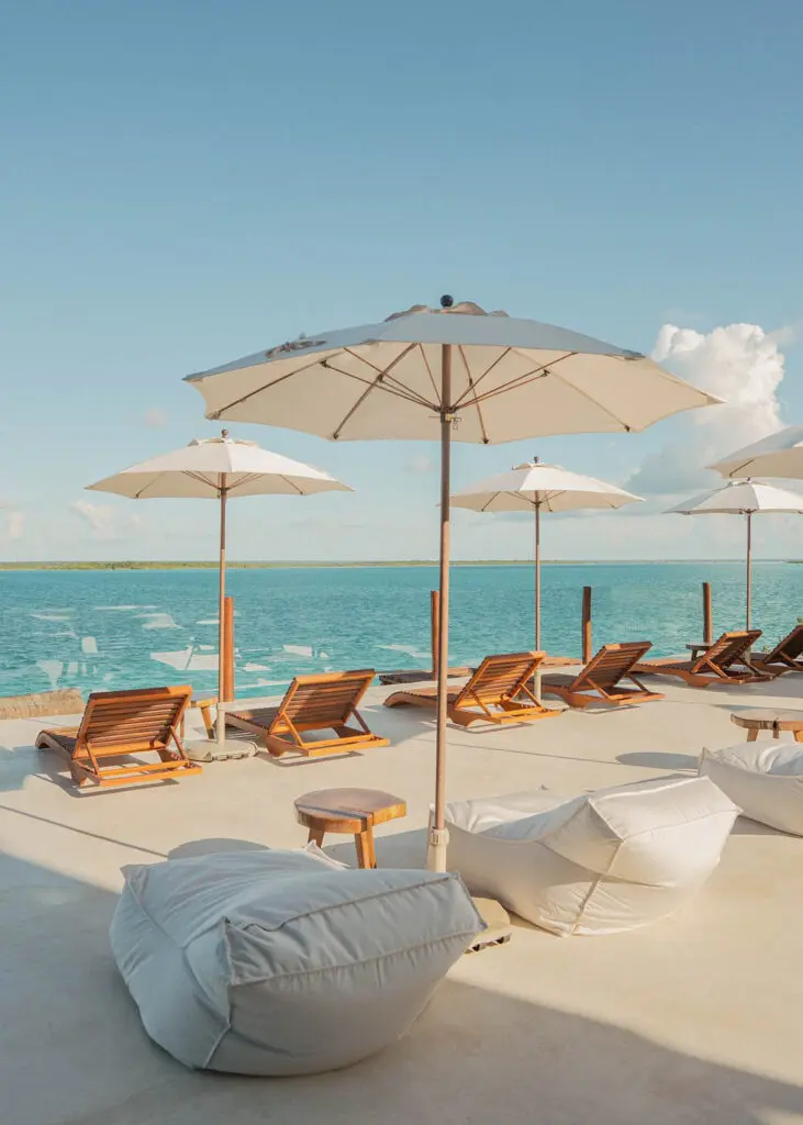 Wooden lounge chairs and white sun loungers with white umbrellas line a cement deck overlooking Bacalar lagoon at Puerta del Cielo Hotel Origen, Bacalar's highest rated waterfront hotel.