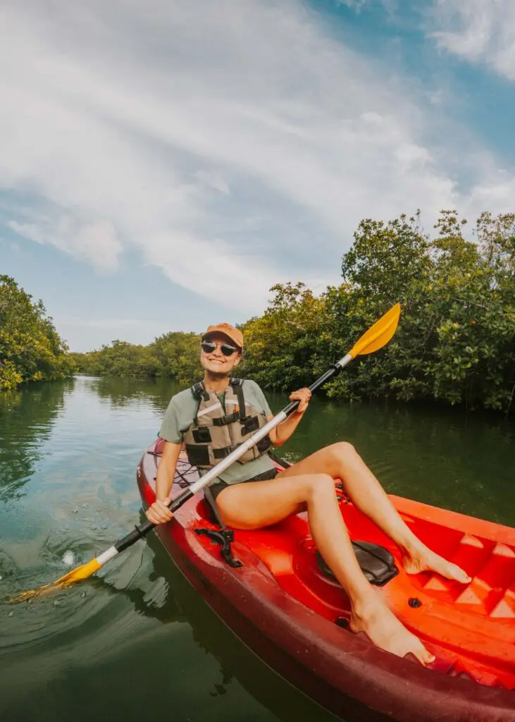 A person wearing a cap, sunglasses, and a life vest is kayaking on a calm, green waterway surrounded by lush greenery and trees. The sky is partly cloudy, reminiscent of a peaceful start to an adventurous Cancun itinerary. The person appears to be enjoying the sunny day.