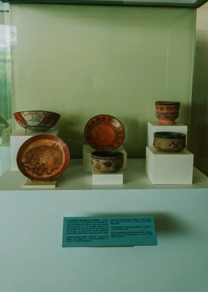 A display case with six ancient pottery pieces on white pedestals, featuring bowls and cups adorned with intricate patterns and earthy tones. As you plan your Cancun itinerary, don't miss this cultural gem. A blue information plaque with text graces the front of the case.