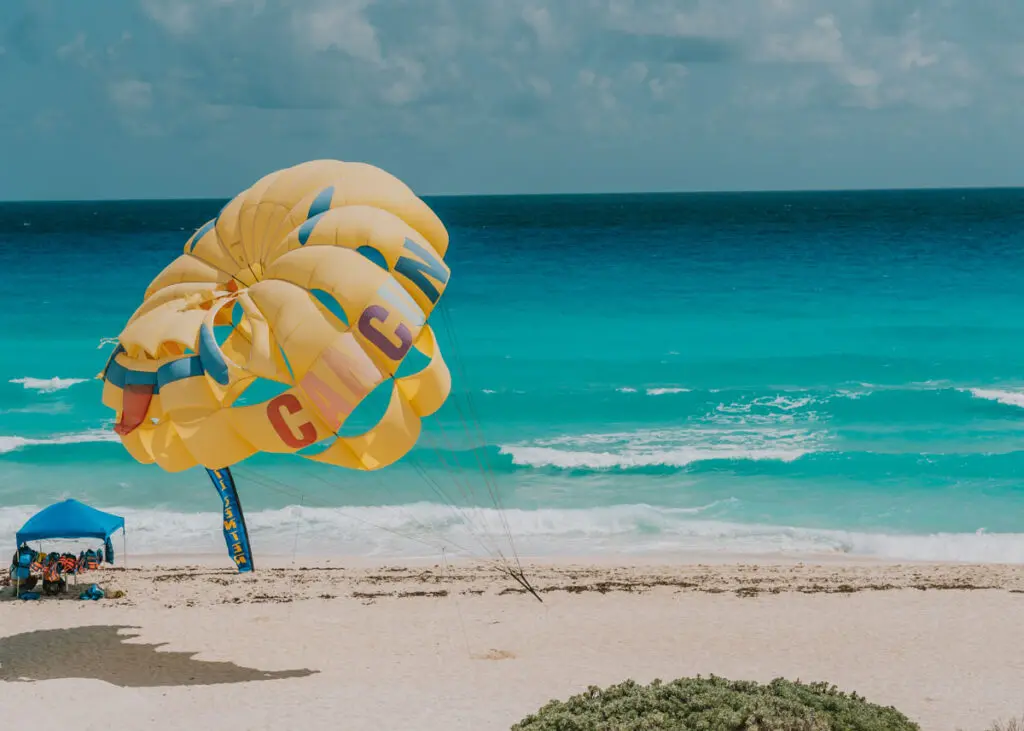 A yellow parasail with "CANCUN" written on it descends toward Playa Delfines, a sandy beach by turquoise ocean waves. A small blue tent shelters people beneath the clear blue sky, perfectly capping off their Cancun itinerary.