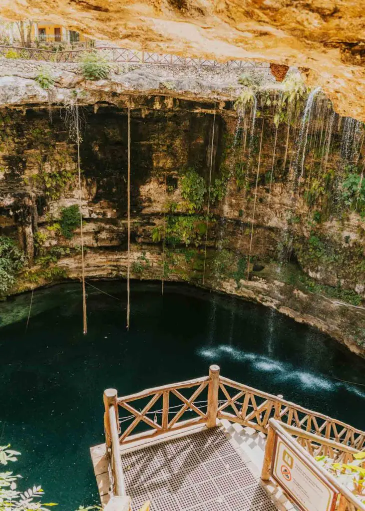 A deep natural sinkhole with clear, dark waters is surrounded by rocky, moss-covered walls and lush greenery. As part of your Cancun itinerary, a wooden walkway with railings descends into the cenote, where thin water streams flow down the walls. A small sign and plants welcome you at the entrance.