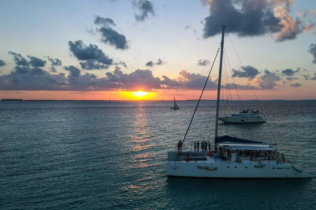 A sailboat with people on deck glides over calm water at sunset. The sky, filled with clouds, casts vibrant colors while another boat in the distance partially obscures the sun. This sunset cruise is a must if you have 3 days in Cancun!