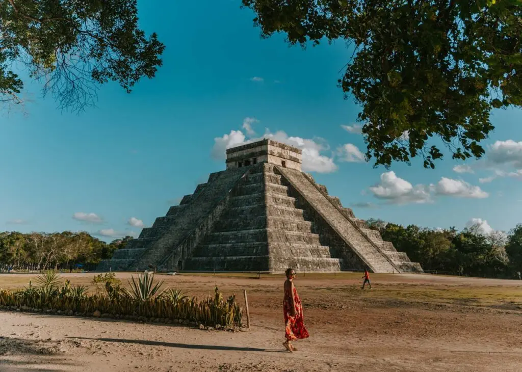 A person in a patterned dress walks near the ancient stone pyramid of Chichén Itzá under a clear blue sky. Trees and fellow travelers offer company, making it a highlight on any Cancun itinerary.