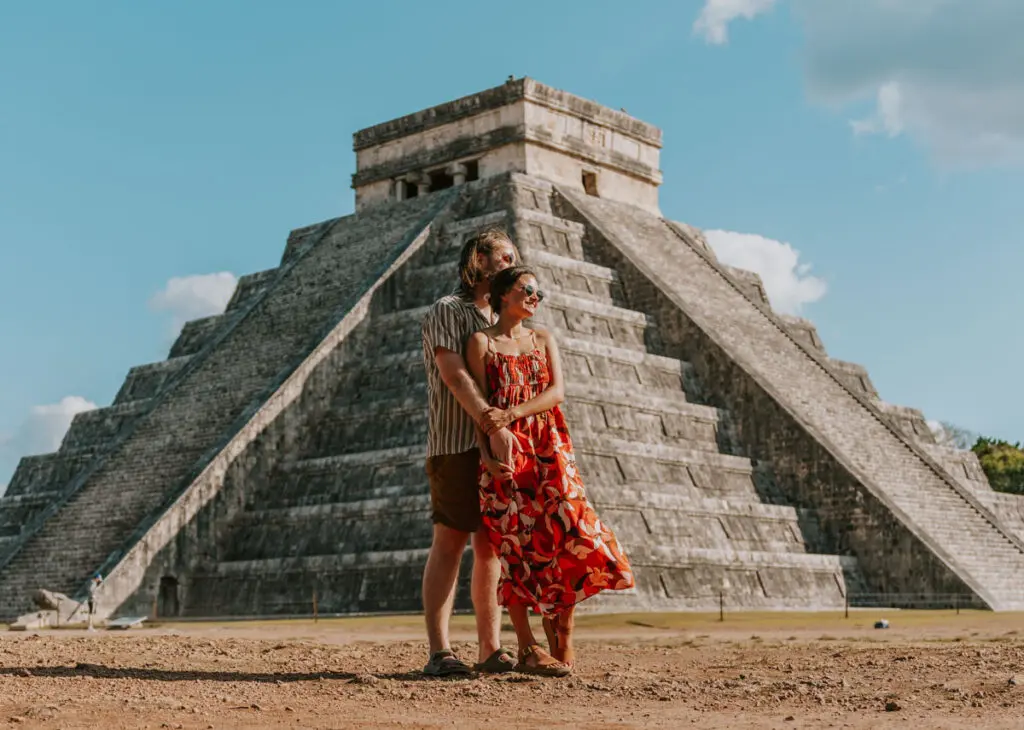 A couple stands smiling in front of a large stone pyramid under the blue sky with clouds at Chichen Itza near Cancun Mexico. The man wears a striped shirt and shorts, while the woman flaunts a colorful dress—perfect highlights for their Cancun itinerary.
