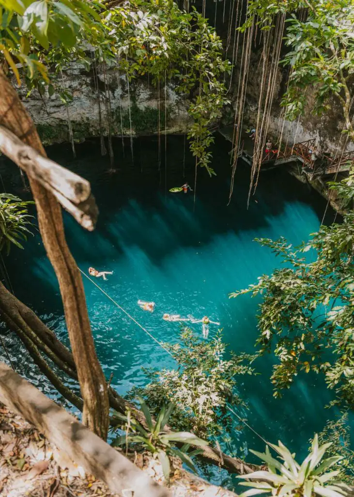 Aerial view of a turquoise cenote surrounded by lush greenery. People swim and float, enjoying the natural pool in this serene setting—a perfect spot on your Cancun itinerary. Long vines hang from the rocks above, enhancing the picturesque scene.