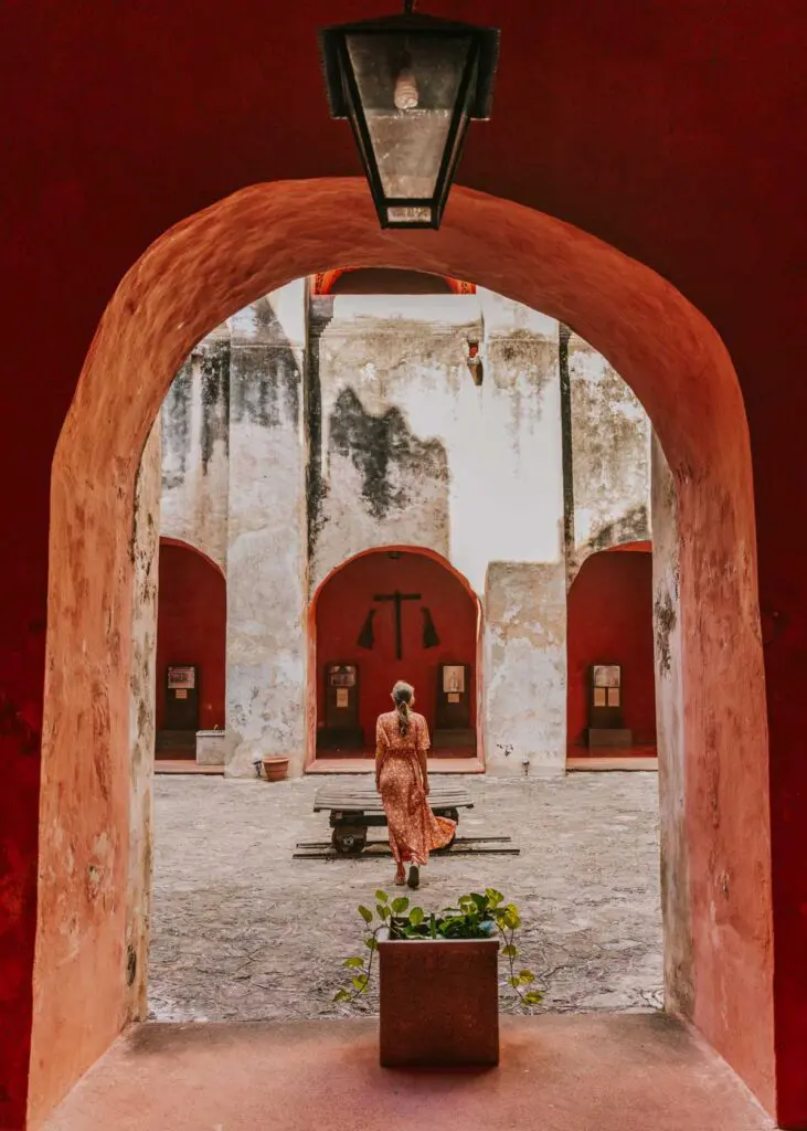 A woman in a long dress strolls through an archway into a courtyard with red and white walls. A lantern hangs overhead, and a small potted plant graces the foreground.