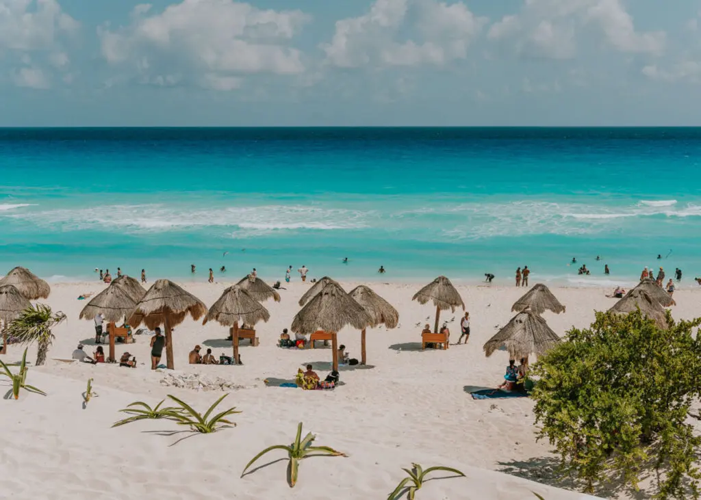 A sunny Playa Delfines scene with turquoise waters and white sand. People relax under straw umbrellas and swim in the sea. Green shrubs and plants dot the foreground, enhancing your Cancun itinerary. Fluffy white clouds scatter across the vibrant blue sky.