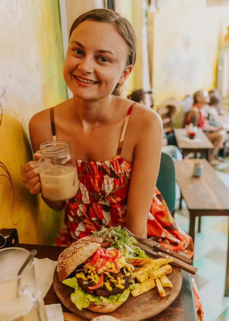 A woman in a colorful dress smiles at a table, holding a jar of iced coffee. Her plate boasts a loaded burger and fries, garnished with sprouts.
