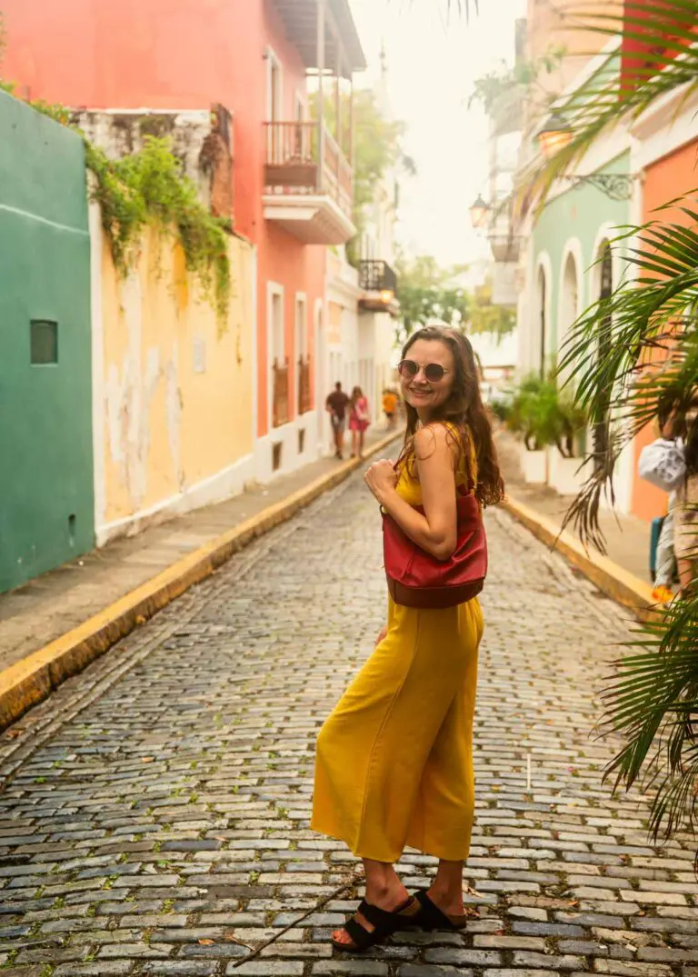 Woman in yellow set peeks back at the camera while exploring a cobblestone street in Old San Juan, Puerto Rico lined with pastel painted buildings.
