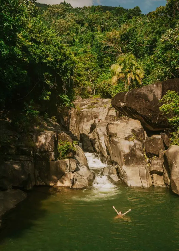 Woman swimming with arms in the air beneath a small waterfall at Charco El Hippie, one of the best places to visit near Rio Grande, Puerto Rico