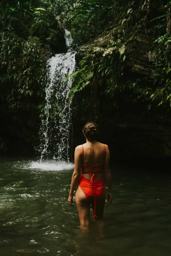 A woman in a red bathing suit wading in a natural pool beneath Juan Diego Falls in El Yunque National Forest, which is one of the best places on this 4 day Puerto Rico itinerary!
