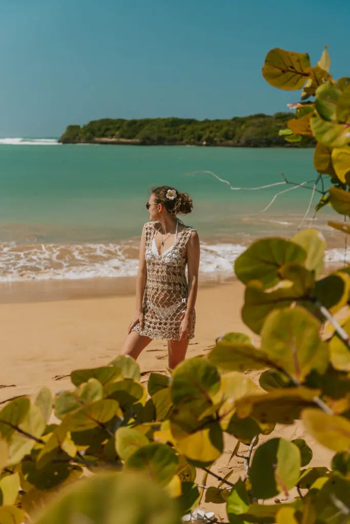 Woman wearing a white bathing suit and crochet coverup stands in front of foliage on a golden sand beach at Playa Piñones in Loiza, Puerto Rico, near Rio Grande town