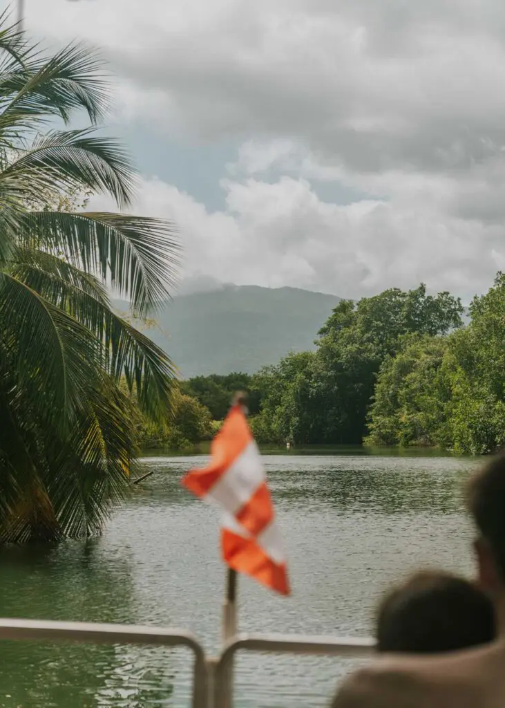 A Puerto Rico flag waves at the front of a pontoon boat ride along Puerto Rico's foliage-lined Rio Espiritu Santo with mountains of El Yunque in the background. 