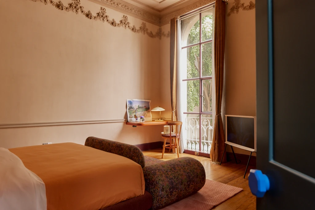 In the artsy Roma neighborhood, this stylish bedroom boasts a patterned sofa, orange bedding, and a large window with a view of greenery. A desk with a lamp and chair is by the window, while modern decor and ornate trim enhance the elegant atmosphere reminiscent of restored mansions.