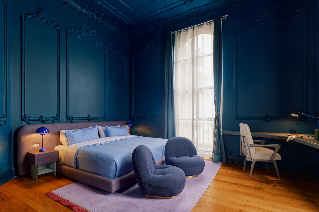 A luxurious bedroom in a restored mansion boasts dark blue walls and ornate moldings. The space features a large bed with blue bedding, two modern lounge chairs, a desk, and a lamp. A large window with sheer curtains invites natural light onto the wooden flooring, creating an artsy boutique hotel vibe.