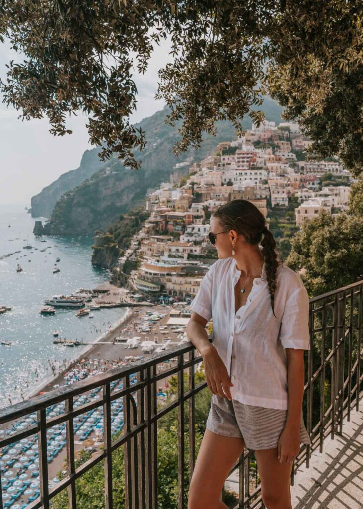 A woman stands by a railing, looking out at the pastel-colored hillside homes and beach umbrellas of Positano on the Amalfi Coast.