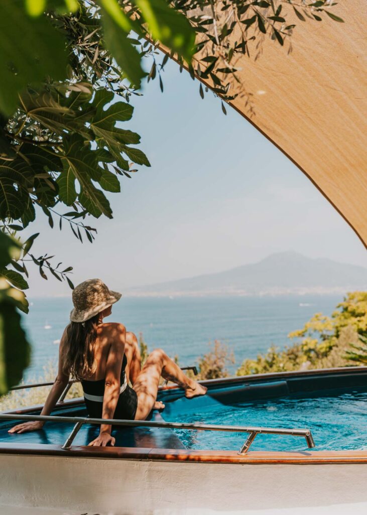 A woman in a black swimsuit and straw hat lounges on the edge of a boat-shaped pool, gazing out at the sea and Mt Vesuvius.
