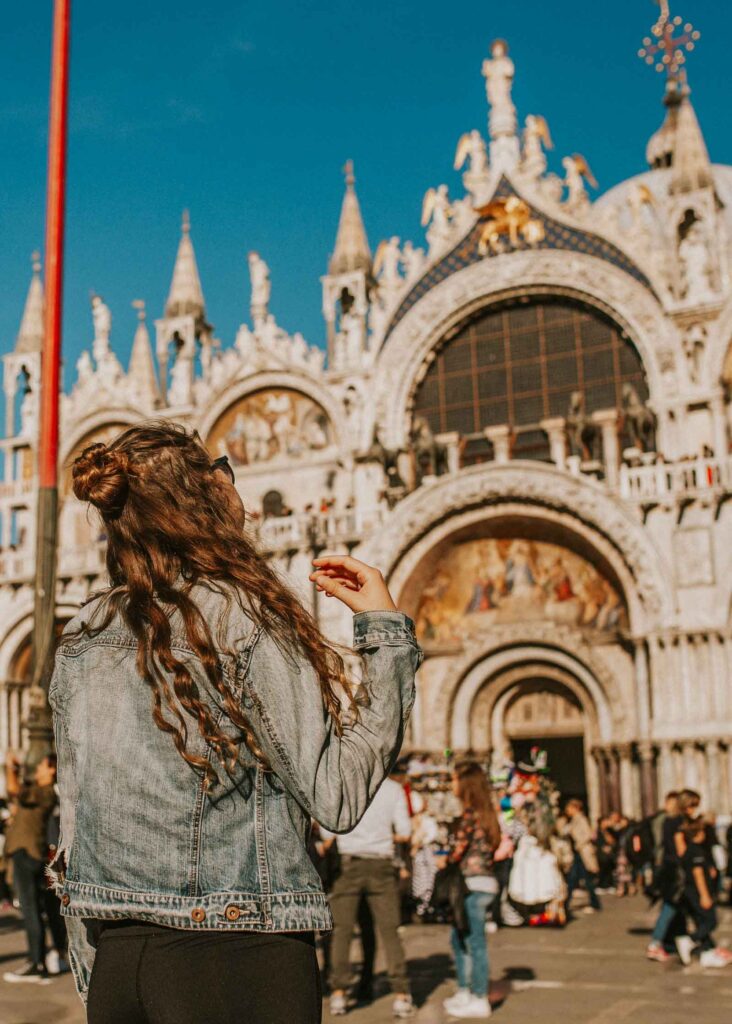 A woman with long wavy brown hair wearing a denim jacket and black pants stands in front of the ornate façade of St. Mark’s Basilica in Venice, Italy. 